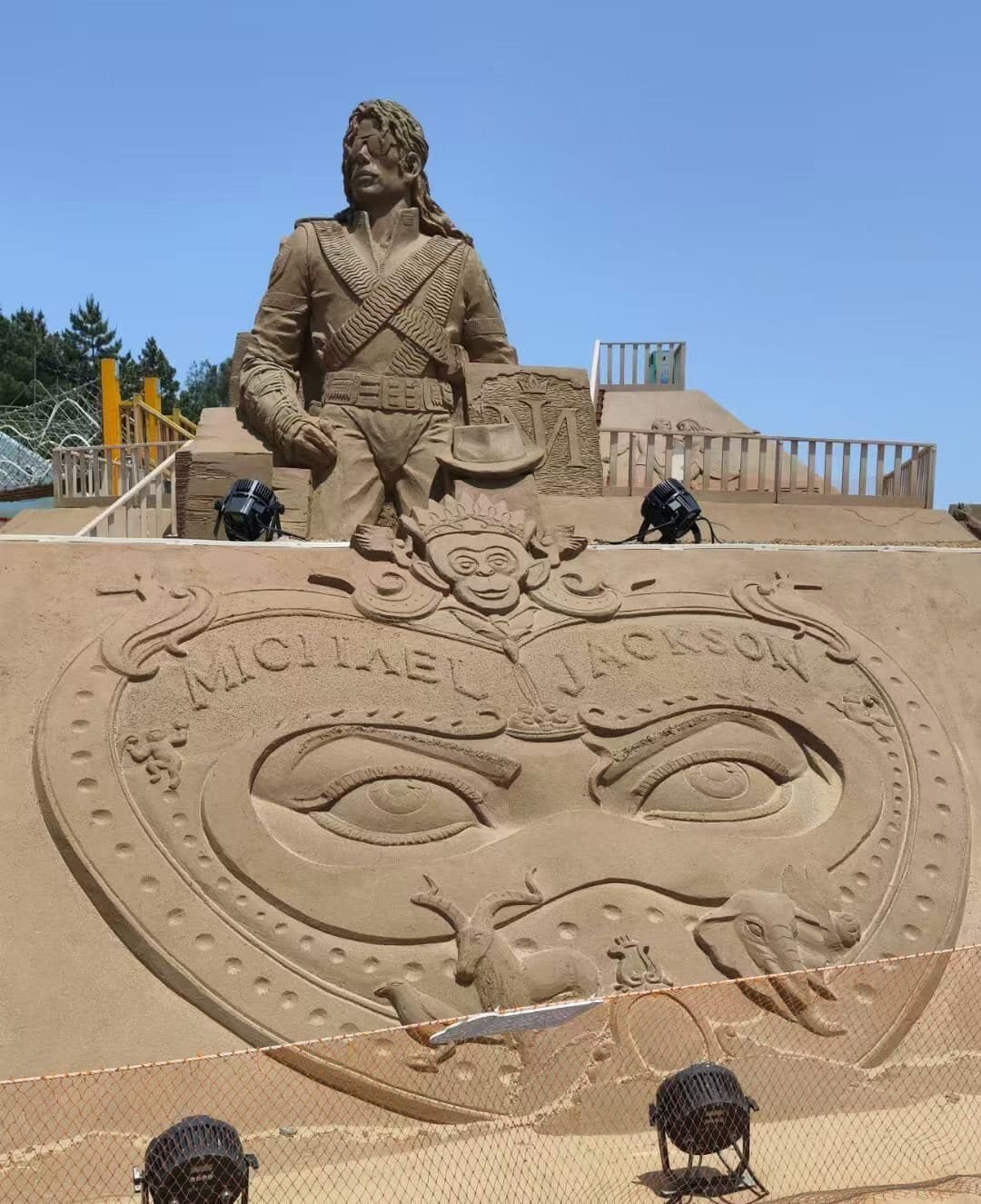 Escultura de areia de Michael Jackson com uniforme militar da era HIStory, em frente a um relevo com olhos, animais e o nome 'Michael Jackson'.