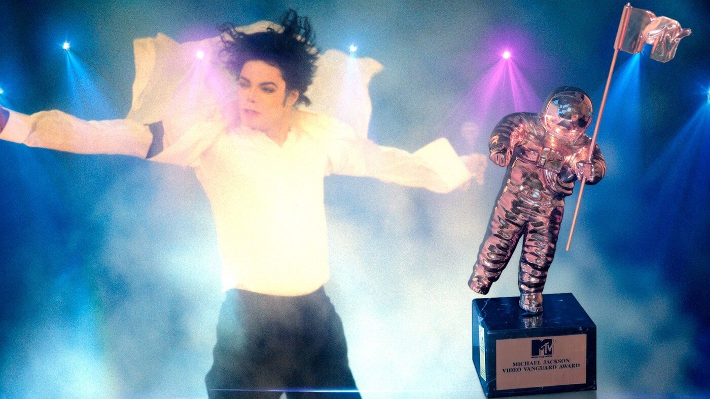 Montagem mostrando Michael Jackson em uma performance com luzes coloridas ao fundo, ao lado da estatueta do astronauta da MTV segurando a bandeira, com a base identificada como “Michael Jackson Video Vanguard Award”.
