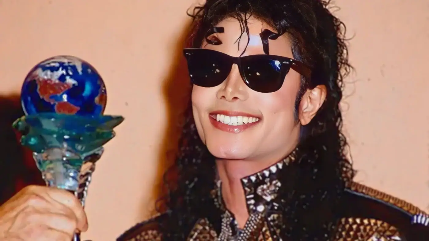 Michael Jackson sorridente, usando óculos escuros e jaqueta preta detalhada em dourado, segura o troféu do Good Scout Humanitarian Award em formato de globo terrestre colorido sobre uma base de cristal.